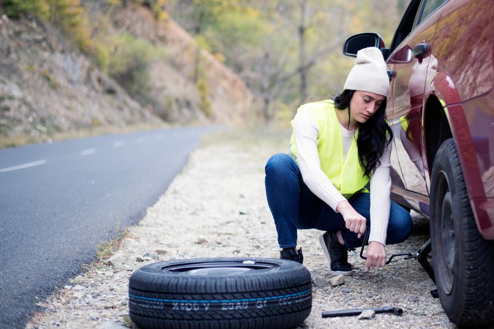 Žena u silnice zvedá odstavené auto pomocí heveru, aby mohla vyměnit poškozenou pneumatiku za rezervu